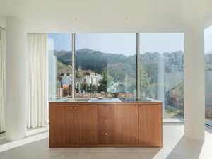 Modern, minimalist kitchen featuring wooden cabinetry and a large window overlooking trees and hills.