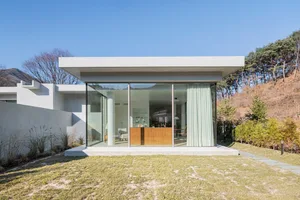 A minimalist modern house with large glass walls and a concrete base on a green hillside.