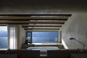 A modern bedroom with concrete walls and a large window overlooking the sea.