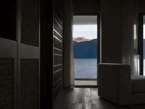 A cozy interior space with wooden floors and glass doors overlooking a stunning sunset over mountains and a body of water.