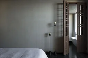 Minimalist bedroom with a white bed, candle holders, and a bathtub in the background, showcasing a modern and serene atmosphe