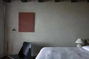 A minimalist bedroom featuring a white sheet on a bed with a black chair, a lamp, and a framed abstract painting on the wall.