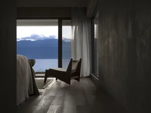 A minimalist bedroom with a view of mountains and water through large windows, featuring a wooden chair and natural lighting.