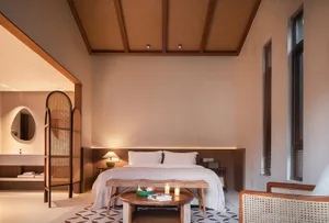 A minimalist bedroom featuring a large bed with a wooden headboard, a wooden bedside table with a lamp, and a wooden armchair