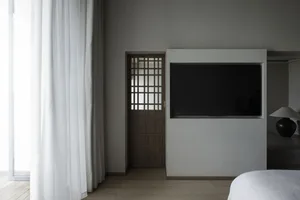 A minimalist bedroom with a wooden door featuring a geometric pattern and a flat-screen TV mounted on a white wall.