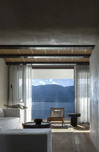 Minimalist living room featuring glass walls with a view of mountains and water, furnished with a white sofa, an armchair, an