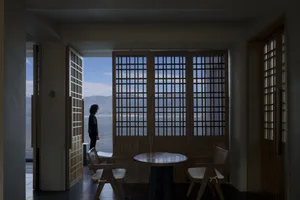 A minimalist interior featuring a view through a window with wooden doors and a table and chairs