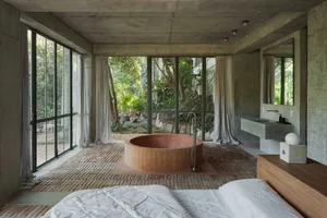 A minimalist bedroom with a concrete and brick floor, a round wooden bathtub, and large windows overlooking greenery.