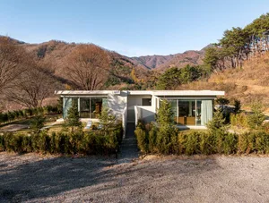 A modern residential house with large glass windows and a concrete exterior surrounded by a landscaped garden with trees and 
