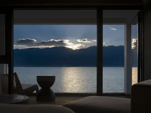 A serene view from a window overlooking the ocean at sunset, with mountains in the background and a wooden chair inside the r
