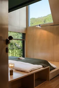 Simple, minimalist bedroom featuring a wooden platform bed with a green blanket and a book on top, large windows with a view
