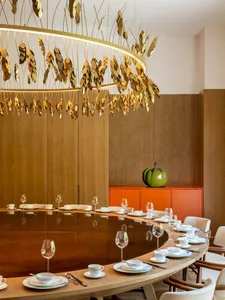 A modern restaurant interior with wooden furniture, featuring a large round table set for dining, a golden chandelier hanging