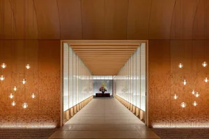 A modern corridor with wooden walls, glass walls, and hanging light bulbs leading to a flower arrangement at the end.