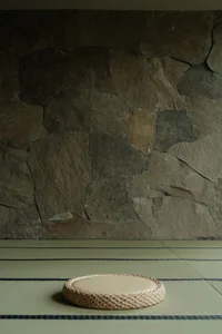 A minimalist floor space featuring a cotton and rattan cushion on tatami matting with a stone wall background.