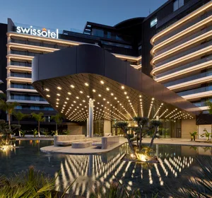 A modern hotel building with a curved glass facade illuminated at night, featuring a water feature and landscaping.