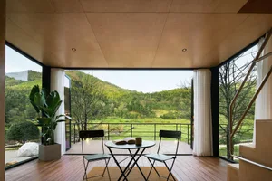 A modern minimalist porch with wooden flooring and curtains overlooking green hills.