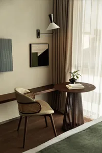 A modern minimalist bedroom featuring a wooden table with a plant and books, a comfortable chair, and a sleek desk lamp.