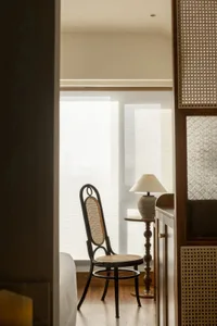 A minimalist residential interior featuring a wooden chair with wicker seat and back, placed near blinds and a lamp on a wood