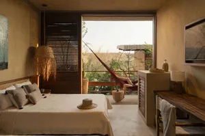 A minimalist bedroom with a bed, bedside table, and a hammock on the balcony overlooking trees.