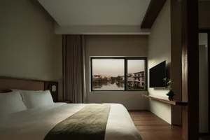 A minimalist bedroom featuring a comfortable bed and a window overlooking a serene waterway and residential buildings.
