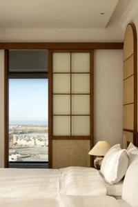 A modern minimalist bedroom with a large window overlooking a cityscape, featuring neutral tones and a wooden headboard with