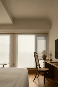 A modern minimalist bedroom featuring a bed with a white sheet, a wooden chair with a woven backrest, a lamp, and a televisio