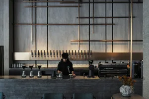 A minimalist coffee bar featuring a concrete counter with a person serving coffee, industrial-style lighting fixtures hanging