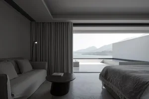 Modern minimalist bedroom featuring a large window with a view of mountains, a bed with a textured blanket, a couch, and a sm