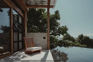 Minimalist outdoor lounge area with a wooden chair on a terrace overlooking greenery and a pool.