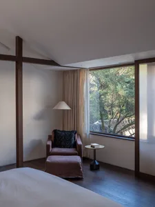A minimalist bedroom featuring a taupe-colored armchair and footstool next to a window with natural light streaming in, set a