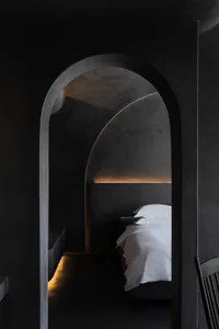 A minimalist bedroom with a concrete headboard and floor, featuring a single white bed and soft lighting.