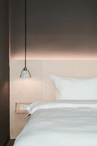 A minimalist bedroom featuring a white bed and wooden walls with a pendant light hanging above.