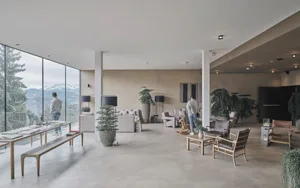 A modern minimalist lobby featuring concrete flooring and walls, with seating areas, potted plants, and a view of mountains t