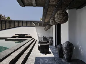 A modern minimalist outdoor living area featuring concrete and stone surfaces, rattan pendant lights, and a poolside seating 