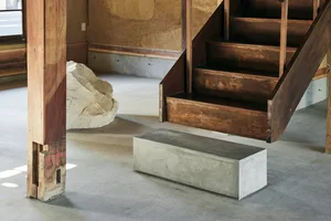 A photograph depicting an interior architectural design featuring wooden stairs, a concrete bench, and a large rock.