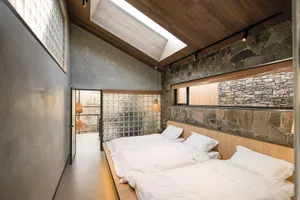 A modern minimalist bedroom with concrete and stone walls, featuring three beds and natural light from a skylight and windows