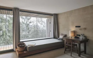 Modern bedroom with a floating bench, wooden beams, and a view of the forest through large windows.