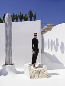 A person wearing a black outfit poses on a marble platform in a modern architectural setting with cacti and wicker baskets.