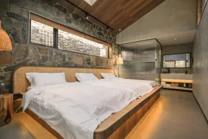 Modern minimalist bedroom with white sheets, wooden headboard, and stone accent wall.