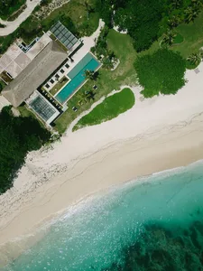 Aerial view of a modern studio house by the beach with a long pool, surrounded by greenery and sand.