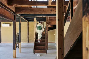 A woman dressed in traditional Japanese attire climbs a wooden staircase in a minimalist, residential setting.