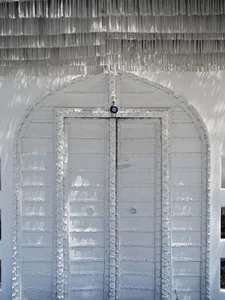 A white metal door with a decorative fringe on top, set against white walls with sunlight casting shadows.