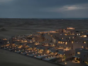 Aerial view of a luxurious desert resort during nighttime with sand dunes in the background and illuminated buildings.
