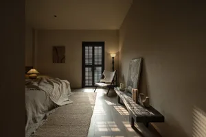 A minimalist bedroom with a bed covered in a textured blanket, a modern chair, and a wooden bench against a light-colored wal