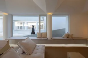 A minimalist living room with neutral-toned couches and cushions, large windows offering a view of an exterior space.