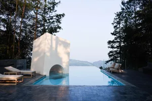 A minimalist concrete swimming pool with a cube-shaped structure and an infinity edge, surrounded by trees and overlooking di