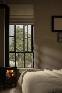 A cozy bedroom with a wooden fireplace and large window overlooking green trees.