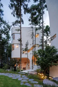 A modern contemporary house with white exterior design and large glass windows surrounded by tall trees.