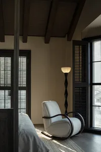 A serene bedroom interior featuring a white upholstered rocking chair, a floor lamp, and a bed with neutral-toned bedding and