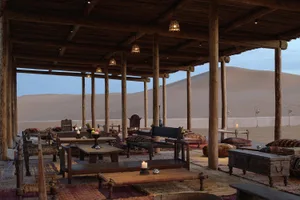 An indoor desert camp with wooden structures, wooden tables and chairs, and a view of a sand dune.